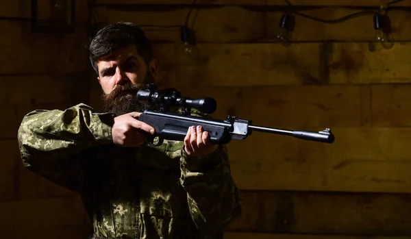 Man with beard wears camouflage clothing, wooden interior background. Hunter, brutal hipster with gun aiming before hunting, copy space. Huntsman concept. Macho on strict face at gamekeepers house — Stock Photo, Image