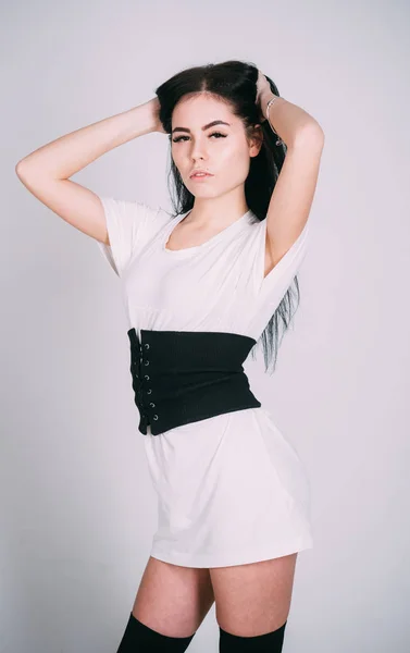 Fille brune porte une robe à la mode et des bas. Concept de tenue à la mode. Dame en robe élégante avec une large ceinture noire à la taille. Femme sur mystérieux visage coûteux posant, mur blanc sur fond — Photo