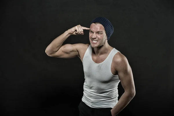 Man with muscular arms pointing at head with index finger. Masculinity concept. Macho on aggressive face with strong muscles look brutal, black background. Man in hat and sloven sleeveless undershirt