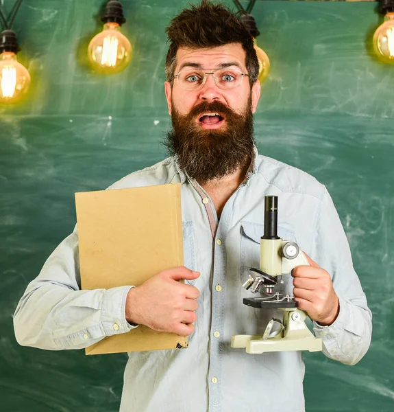 戴眼镜的老师手持书本和显微镜。科学概念。科学家在背景上拿着书和显微镜, 黑板。人与胡子和胡子在惊奇的面孔在教室里 — 图库照片