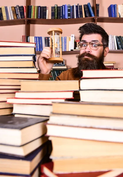 Homem no rosto surpreso detém ampulheta enquanto estuda, estantes de livros no fundo. Conceito de fluxo de tempo. Professor ou estudante com barba estudando na biblioteca. Homem, cientista de óculos olha para ampulheta — Fotografia de Stock