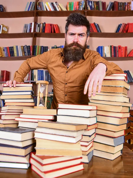 Concept de bibliothécaire. L'enseignant ou l'élève avec la barbe se tient à table avec des livres, déconcentré. Homme au visage réfléchi se tient entre des piles de livres, tout en étudiant à la bibliothèque, étagères sur fond — Photo