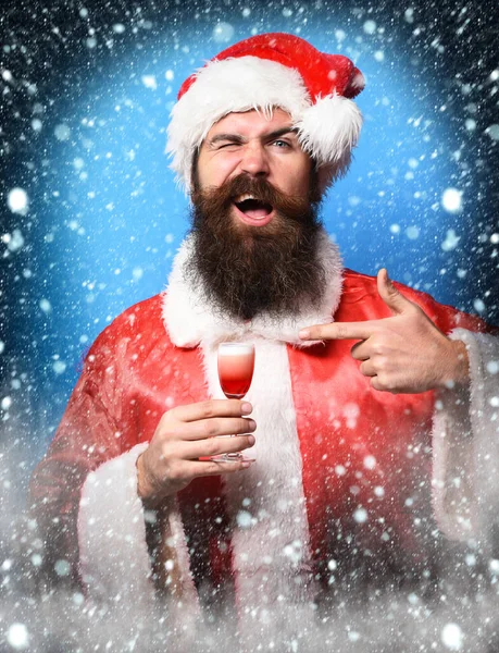 Pohledný vousatý Santa Claus muž s dlouhým vousem na usmívající se tváři drží sklo alkoholické střely v červené Vánoce nebo vánoční svetr a Nový rok klobouk na modrém pozadí studia — Stock fotografie