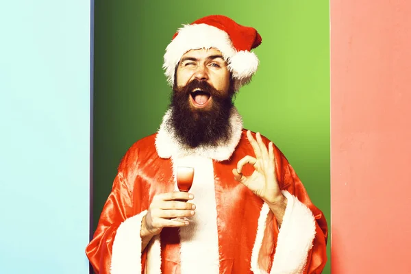 Yakışıklı, sakallı, uzun sakallı, komik sakallı Noel Baba elinde kırmızı noel ya da yılbaşı ceketi ve renkli stüdyo şapkası olan alkolik bardağı tutan adam. — Stok fotoğraf