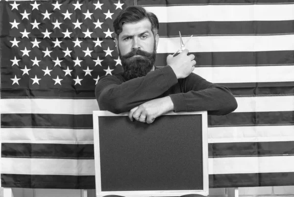 School the best in barbering. Bearded man holding scissors and blackboard in school. Hipster teaching hairdressing on american flag background. Barber training school. Barbershop school, copy space — Stock Photo, Image