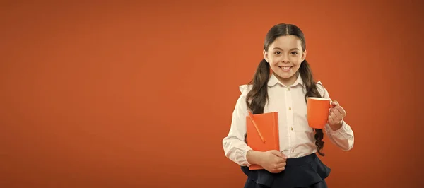 Un sabor saludable. Linda chica con taza de té y cuaderno sobre fondo naranja. Un niño bebiendo té para desayunar. Niño pequeño disfrutando del té de la mañana. Niño tomando un descanso para el té en la escuela, espacio para copiar — Foto de Stock