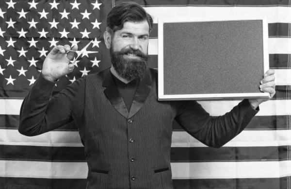Professional man. Man teacher giving lesson on american flag background. Bearded man holding scissors and blackboard in school. Man barber providing knowledge and skills of cutting hair, copy space