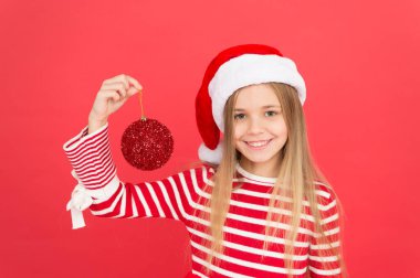 Noel 'i neşeyle süsleyin. Küçük Noel Baba kız Noel ağacı topu tutuyor. Kırmızı parıltılı topuyla mutlu çocuk gülüşü. Noel mücevheri ya da top süsü. Kırılgan cam top süslemesi. Xmas ampulü ya da takısı