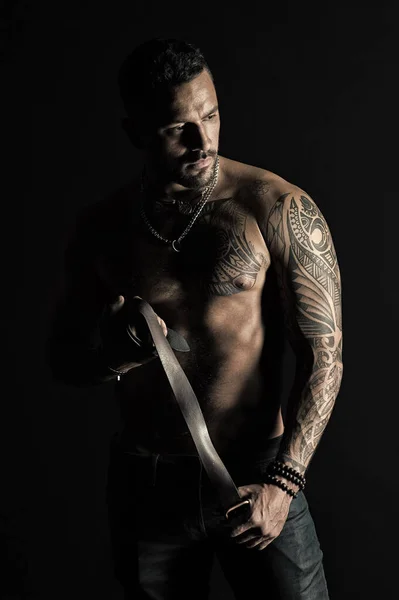 Fashion model with leather belt in jeans. Man with tattoo design on skin. Bearded man with muscular torso. Sportsman with tattooed arm and chest. Bodycare with fitness and sport in vintage filter