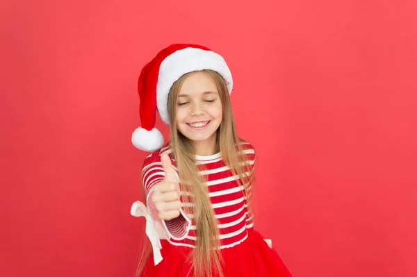 Пальцы вверх к Новому году. Счастливая девочка улыбается с новогодним взглядом. Маленький ребенок показывает большие пальцы вверх жеста красный фон. Новогодняя вечеринка. С Новым годом. Счастливого Рождества! — стоковое фото