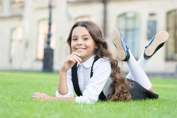 Tillbaka till lektionen och ser glad ut. Glad liten flicka koppla av på grönt gräs. Glada små barn med glada leenden och formell look. Underbara barn njuta av skolan tid. Hon verkar lycklig i skolan. — Stockfoto