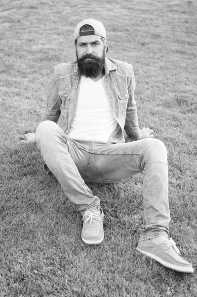 Mantenerlo informal. Hombre barbudo en traje casual y cómodo sentado sobre hierba verde. Hipster en estilo vaquero casual en verano al aire libre. Casual y cómodo — Foto de Stock