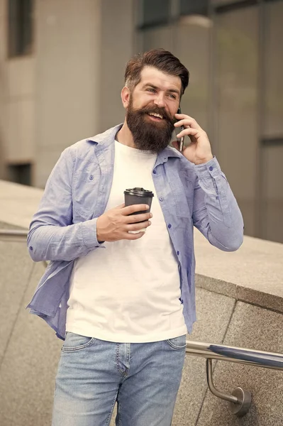 Keep in touch. Man bearded walk with smartphone and coffee cup urban background. Man smiling face smartphone. Guy pleased answer call smartphone. Communication concept. Glad to hear. Good news