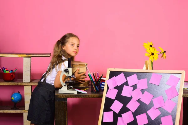 Menina coloca livros na mesa com microscópio e quadro-negro — Fotografia de Stock