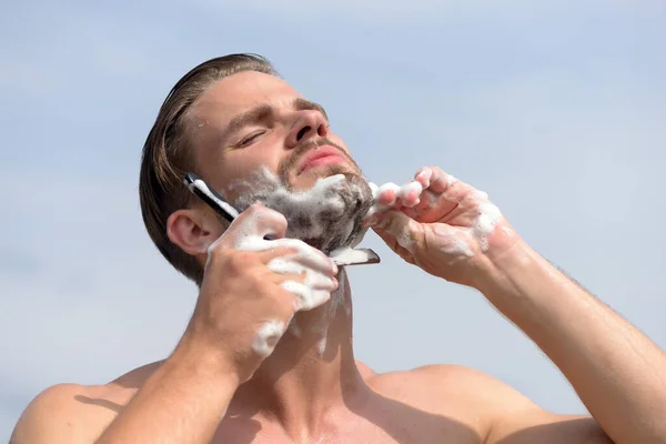 Macho shaving chin with razor on clear sky background — Stock Photo, Image