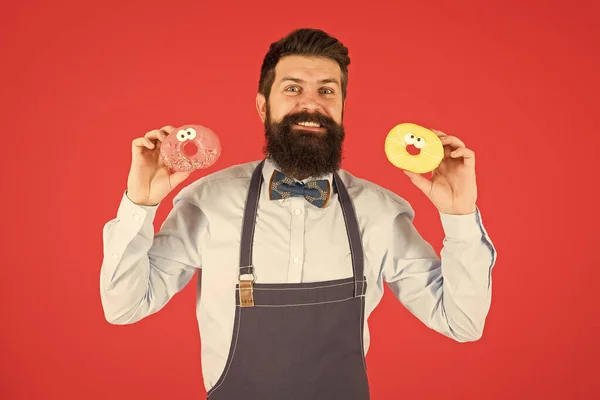 Donuts assados com caras engraçadas. Padeiro feliz segurando lanche caseiro no fundo vermelho. Hipster sorrindo com sobremesa recém-assada e vitrificada. Homem barbudo com donuts cozidos nas mãos — Fotografia de Stock