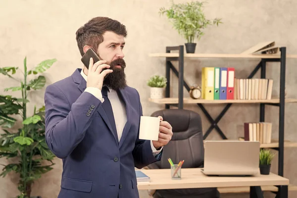Мужчина бородатый бизнесмен держит чашку и смартфон. Кофе залог успешных переговоров. Зависимость от кофеина. Мобильный звонок. Начните день с кофе. Кофе расслабляющий перерыв. Босс наслаждается энергетическим напитком — стоковое фото
