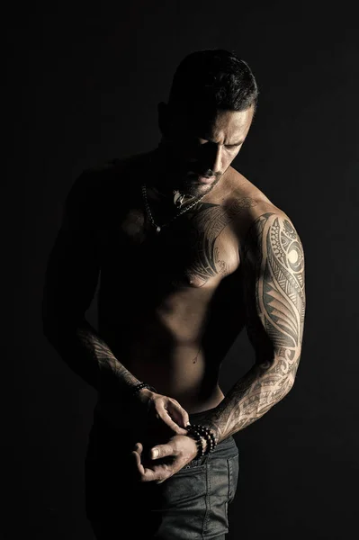 Masculinidad y fuerza. Hombre barbudo con tatuaje en brazo fuerte. Hombre con torso muscular. Moda machista sin camisa con pulseras de cuentas en jeans. Moda con estilo y tendencia, filtro vintage — Foto de Stock