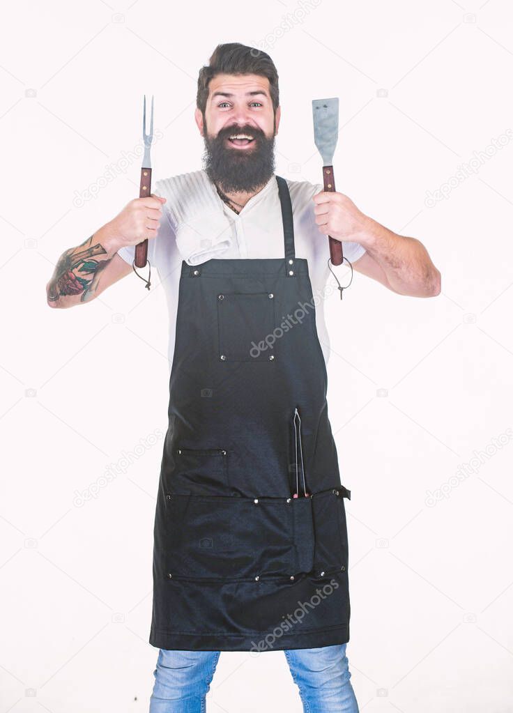 He is a great cook. Happy grill cook with cooking utensils. Bearded man holding fork and spatula for cooking and serving barbecue. Master cook wearing grilling apron. Chief cook in workwear. Cookout
