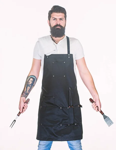 Servir barbecue. Homme barbu avec des outils de barbecue grill dans les mains. Hipster en tablier avec ustensiles métalliques pour barbecue. Cuisinière au barbecue. Préparation de la nourriture sur le gril à l'aide d'un ensemble barbecue — Photo