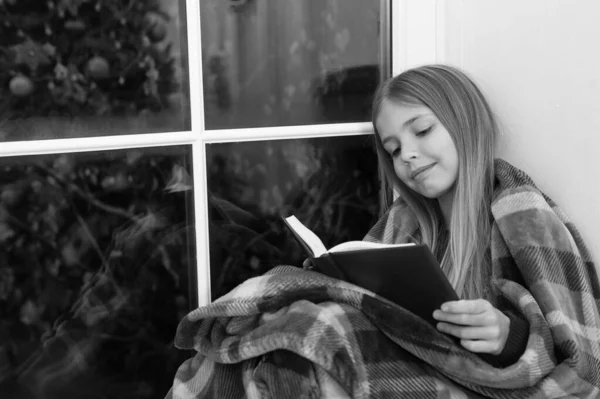 Conte de fées pour des vacances joyeuses. Petit lecteur enveloppé de plaid assis sur le rebord de la fenêtre. Petite fille aime lire l'histoire de Noël. Un petit enfant lit un livre le soir de Noël. Livre pour enfants. Esprit de Noël magique — Photo