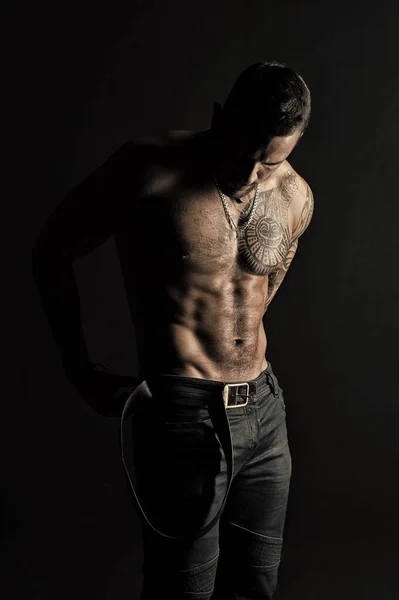 Sportsman with tattooed arm and chest. Bearded man shirtless with fit torso. Man with tattoo design on skin. Fashion model put leather belt on jeans. Bodycare with fitness and sport, vintage filter — Stock Photo, Image