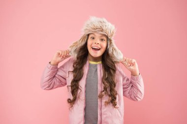 Sıcak bir gülümseme. Mutlu çocuk pembe arka planı. Soğuk mevsim için sıcak kıyafetler. Çocuk modası. Moda kızı hipster 'a benziyor. Sonbahar tarzı. Çocukluk aktivitesi. Tüylü kulaklıklı şapka aksesuarı. Küçük kız kış şapkası