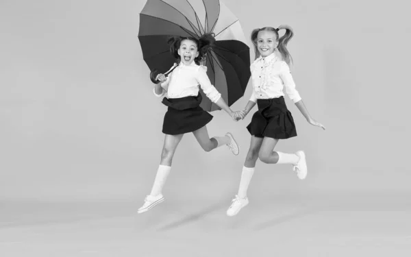 Повернутися до школи в будь-яку погоду. Маленькі дівчата початкового шкільного віку посміхаються під барвистою парасолькою на жовтому тлі. Щасливі маленькі діти поспішають до школи. Насолоджуйтесь шкільними канікулами восени — стокове фото