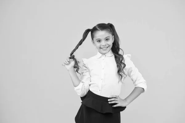 Su peinado retorcido. Pequeña niña enrollando el pelo largo alrededor de su dedo sobre fondo amarillo. Niño pequeño con peinado de cola de caballo en uniforme escolar. Niño con nuevo peinado para volver a la escuela — Foto de Stock