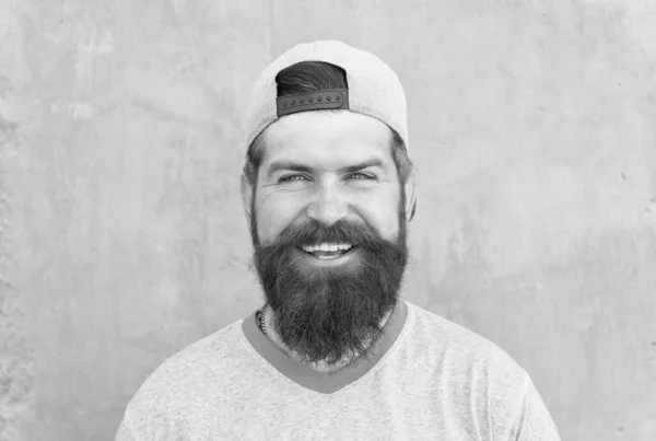 Mantener el cabello sano. Feliz hipster con forma de barba y pelo peinado con gorra de béisbol. Hombre barbudo sonriendo con el pelo de la cara sin afeitar. Tipo peludo con barba elegante y el pelo bigote en la pared gris — Foto de Stock