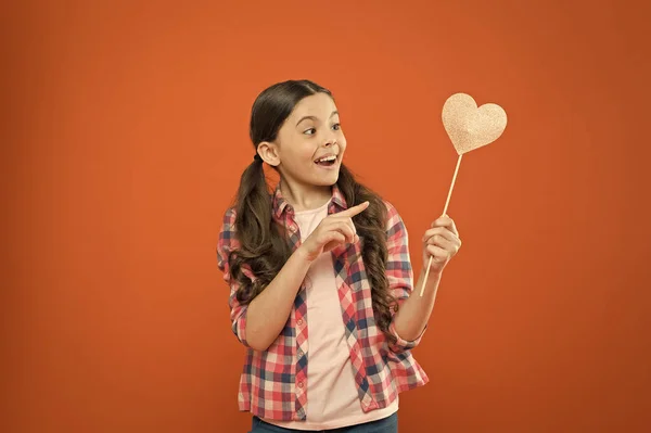 Look at this heart. Cute little child pointing finger at prop heart on orange background. Adorable small girl holding valentines heart on stick. Take my heart and love — Stock Photo, Image