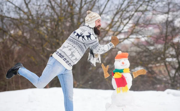Man cheerful bearded hipster knitted hat and warm gloves play with snowman snow outdoors. Have fun winter day. Let it snow. Christmas holidays. Active lifestyle. Snow games. Leisure on fresh air