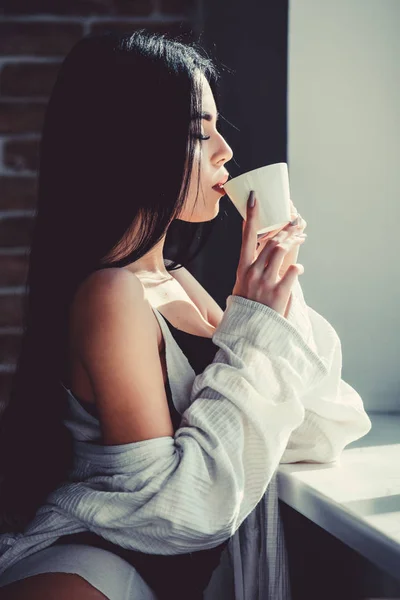 Det bästa kaffet för hennes dagliga rutin. Sensuell flicka dricker hennes favorit morgonkaffe. Söt kvinna dricker färskt varmt kaffe på fönster. Sexig tjej som håller keramiska kaffekopp — Stockfoto