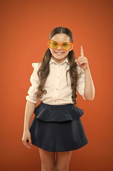 Забавная идея. Маленькая девочка в смешных очках указывает пальцем на оранжевый фон. Забавный ребенок в солнечных очках с цветным фильтром. Мои глаза странные. — стоковое фото