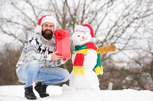 Χειμερινές διακοπές. Ο άνθρωπος έφτιαξε χιονάνθρωπο. Ο 'γιος Βασίλης διασκεδάζει έξω. Χειμερινοί αγώνες. Guy χαρούμενο πρόσωπο χιονισμένο φόντο της φύσης. Hipster με γενειάδα κρατήσει κουτί δώρου. Έννοια έκπληξη. Χειμερινή δραστηριότητα — Φωτογραφία Αρχείου