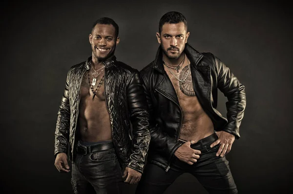 Two male models posing together. Desire and seduction concept. African guy with joyful smile. Hispanic man with geometrical tattoo on hairy chest. Bodybuilders wearing leather jackets on bare torsos — Stock Photo, Image