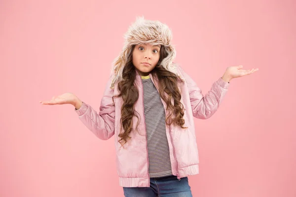 at a loss. warm clothes for cold season. fur hat accessory. small girl winter earflap hat. bewildered child pink background. kid fashion. girl look like hipster. autumn style. Childhood activity