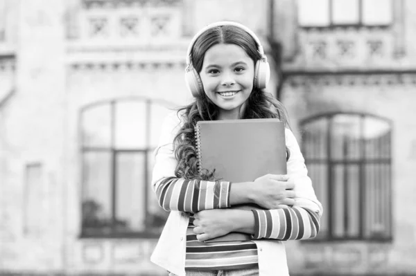 Programas de áudio para alunos. Menina feliz ouvir áudio livro tocando em fones de ouvido. Criança pequena desfrutando de aprendizagem de áudio. Criança em fones de ouvido estéreo segurando materiais de áudio para lição — Fotografia de Stock