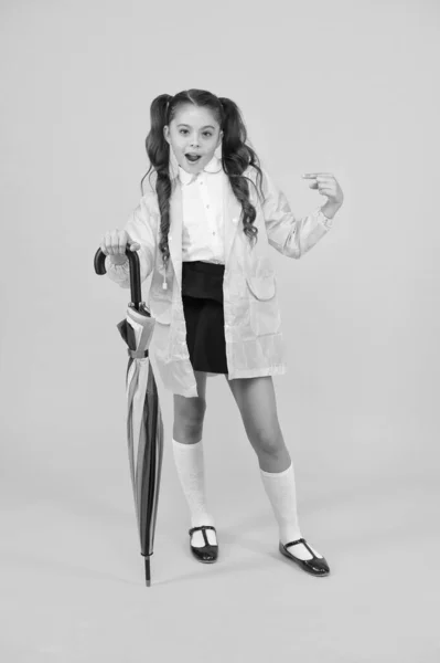 Ze een mode slachtoffer. Schattig school kind wijzend vinger met mode kijken op gele achtergrond. Klein kind in regenjas Holding mode paraplu. Regenkleding en regenachtige dag mode — Stockfoto