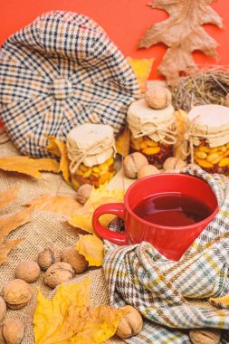 Kupa rahat aromatik çay içecek eşarp ve muamele etmek. Bir fincan çay eşarp kırmızı arka plan düşmüş akçaağaç yaprakları ve ceviz ile çevrili. Rahat sonbahar atmosfer. Sonbahar sezonu kavramı listelemek için