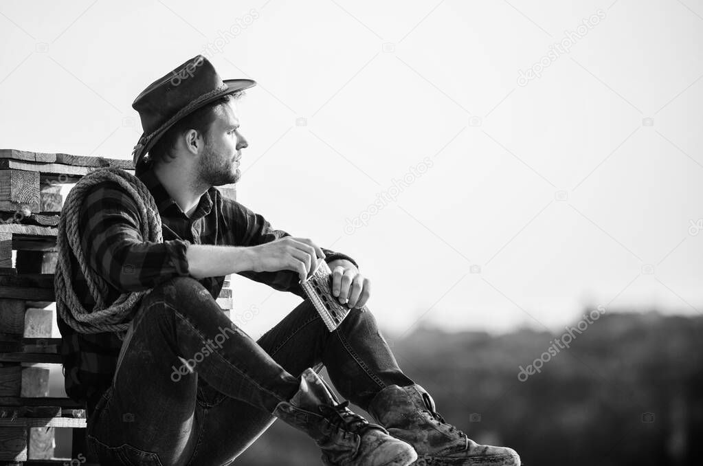 Evening time. Farmer drink alcohol enjoy view from his farm. Watching sunset. Farmer cowboy handsome man relaxing after hard working day at ranch. Romanticism western culture. Farmer in hat sit relax