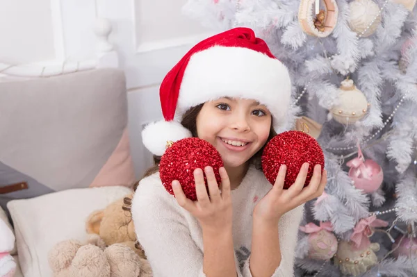 Playful mood. Creating festive atmosphere. Child decorating christmas tree with red ball. Girl kid decorating christmas tree. Kid in santa hat decorating christmas tree. Good vibes. Cheerful kid — Stock Photo, Image