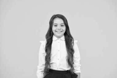 Kızın okulun ilk günü için hazır olması. Mutlu kız okula dönüyor. Sevimli küçük kız sarı arka planda gülümsüyor. Resmi okul üniforması giyen uzun esmer saçlı sevimli küçük kız