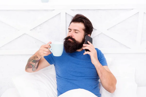 Pleasant conversation. Good morning. Hello dear. Bearded man using mobile technology in bed. Handsome guy talking on phone and drinking coffee at home. Modern life new technology. Technology concept