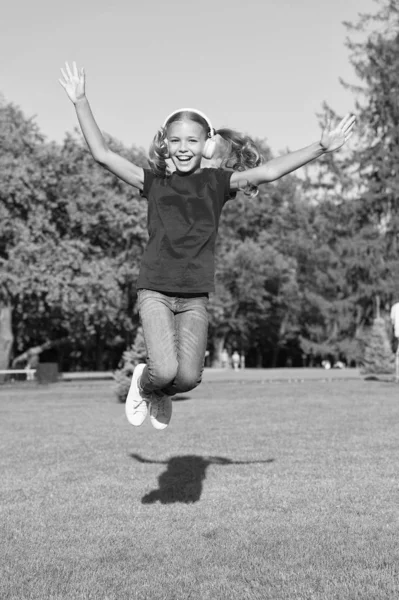 Music of happiness. Happy little child jump high on green grass. Small girl enjoy listening to music in modern headphones. Happiness concept. Feel good. Releasing serotonin dopamine and endorphin