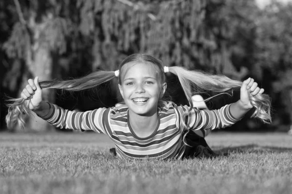 根から先端まで長く健康的です。幸せな小さな女の子は緑の芝生の上で長いポニーテールを保持します。長いブロンドの髪の尾を持つ愛らしい小さな子供の笑顔。髪を長く伸ばしなさい。ヘアケアと治療 — ストック写真