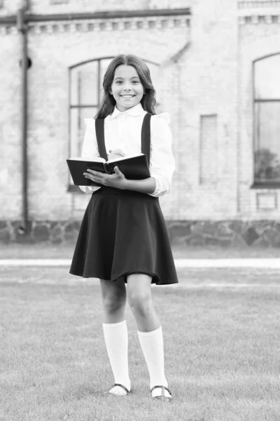 Lección para lector perspicaz. La colegiala feliz toma notas para la lección. Niño pequeño, prepárate para la lección escolar. Adorable niña pequeña mantenga pluma y libro de lecciones. Escuela y educación — Foto de Stock