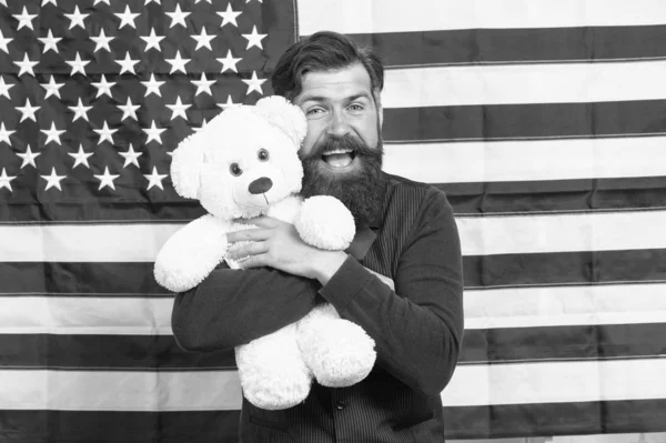 Regalo de juguete navideño. Hombre barbudo sonriendo con regalo de juguete del día de la independencia. Feliz hipster sosteniendo juguete de oso de peluche sobre fondo de bandera americana. Juguete favor para el día de la independencia — Foto de Stock