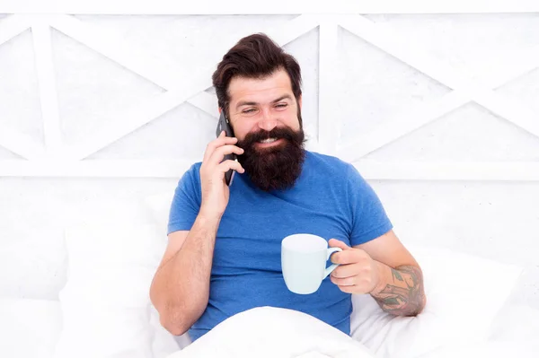 - Доброе утро. Привет, дорогая. Бородатый мужчина, использующий мобильные технологии в постели. Красивый парень разговаривает по телефону и пьет кофе дома. Современная жизнь новые технологии. Технологическая концепция. Приятный разговор — стоковое фото