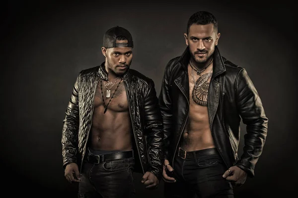 Bikers in leather jackets with brutal look. Bodybuilders with tattoos on their chests. Models with sexy bare torsos, body care and fitness concept. Two men in dark jeans isolated on black background